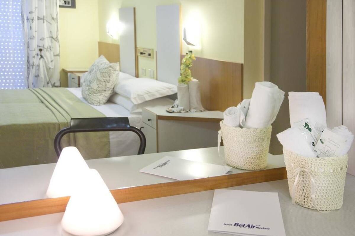 Quarto de hotel com cama, mesa de cabeceira, luminária e espelho. Há duas cestas na mesa, uma com toalhas e outra com um vaso.