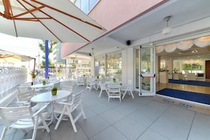 An outdoor seating area with white tables and chairs, a glass door entrance, and a blue carpet leading inside.