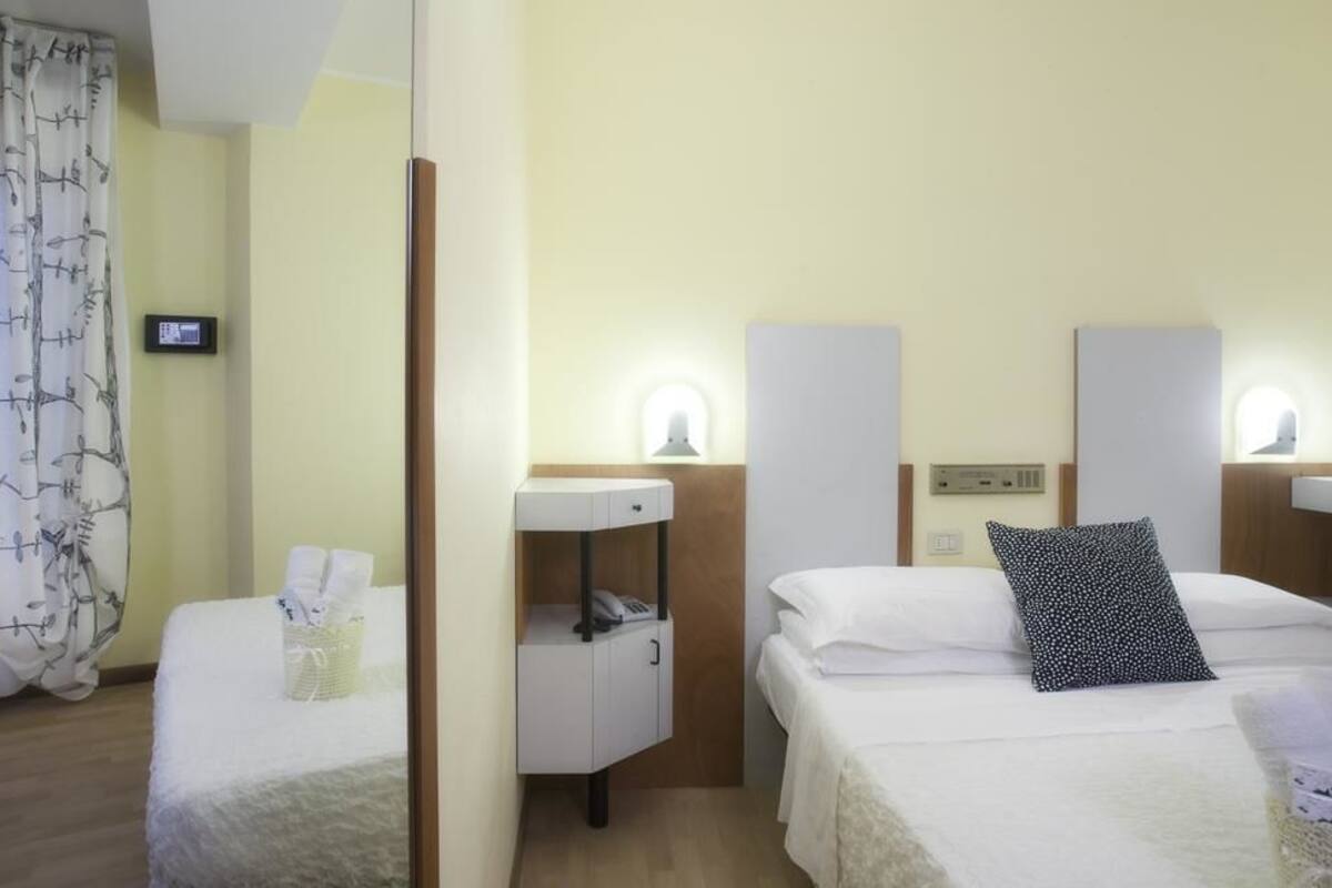 Quarto de hotel com duas camas, uma mesinha de cabeceira com telefone, uma luminária e um pequeno cesto com toalhas.