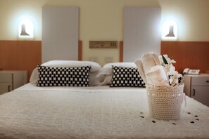 A hotel room with two beds, checkered pillows, a bedside table with a lamp, and a decorative basket with flowers.