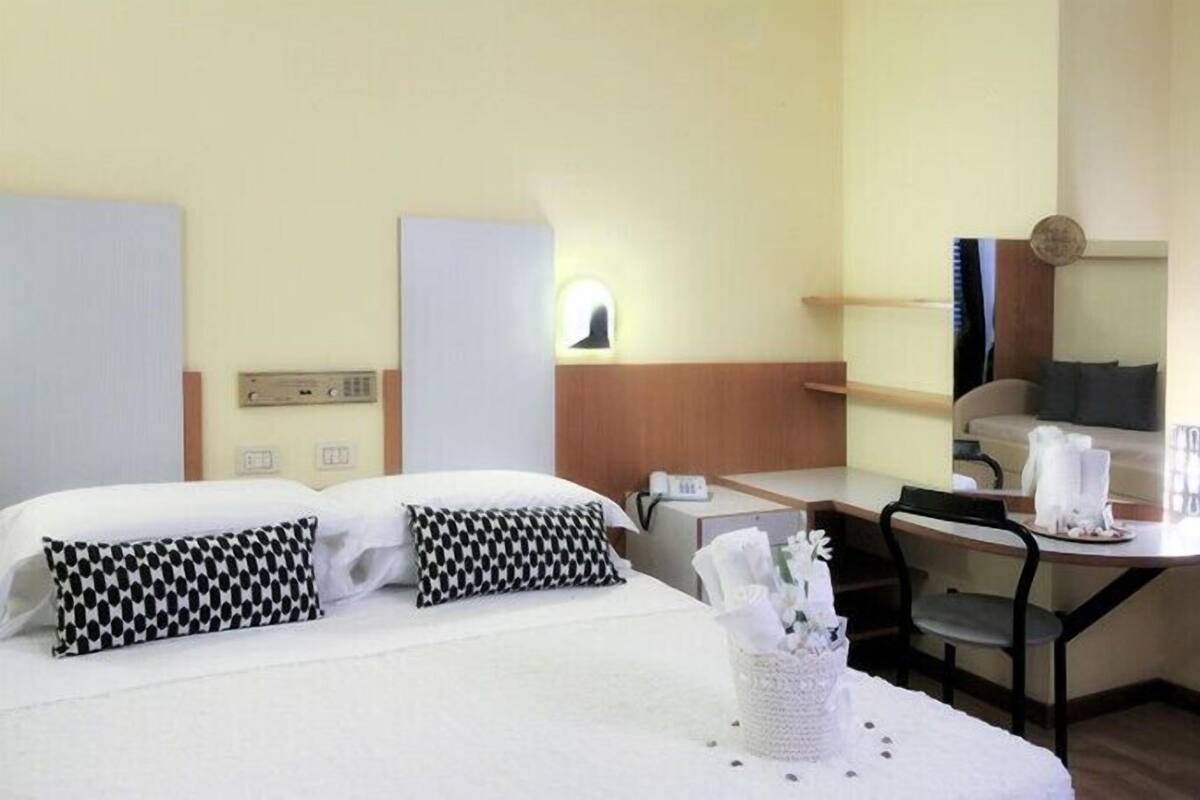 Quarto de hotel com cama, mesa de cabeceira, cadeira, escrivaninha e uma pequena mesa com um vaso de flores.