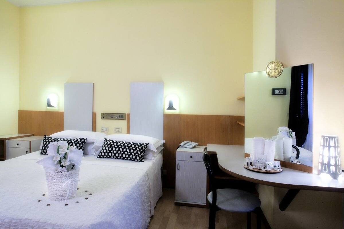 Quarto de hotel com cama, mesas de cabeceira, escrivaninha, cadeira, um pequeno armário e um espelho.