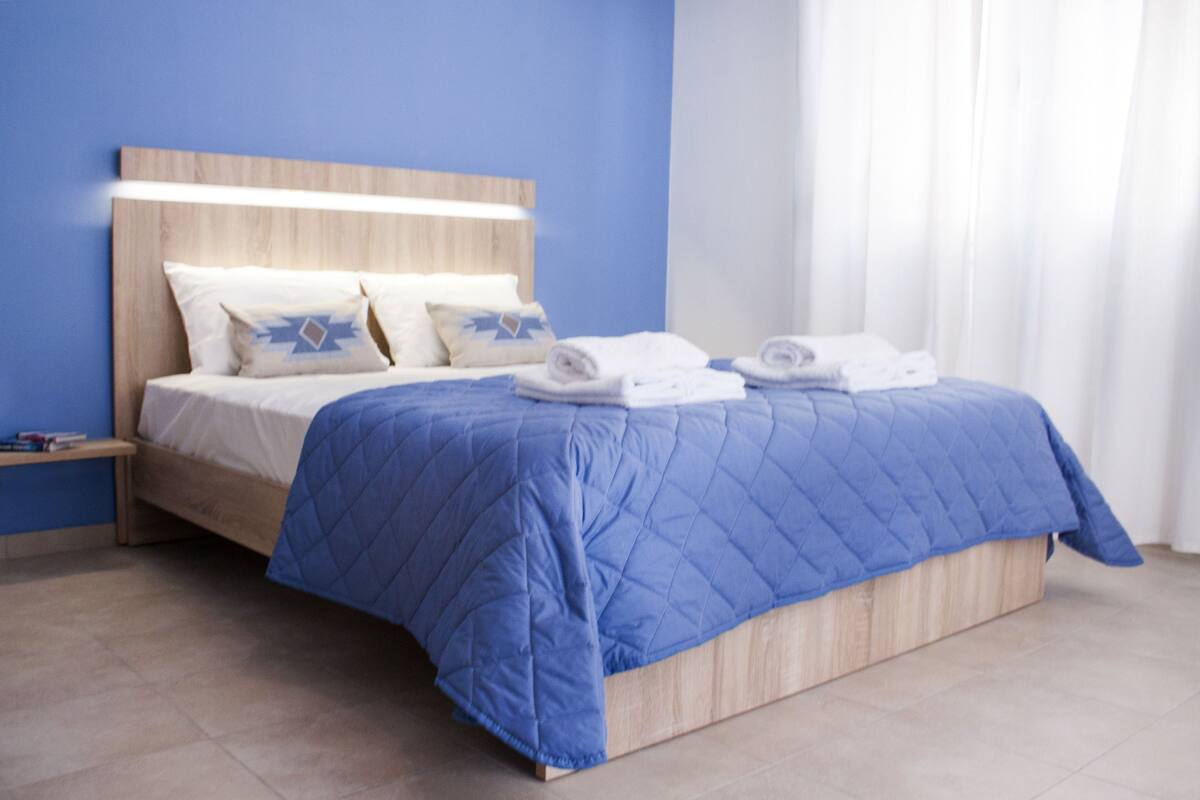 A modern bedroom with a wooden headboard, a blue quilted bedspread, and neatly folded towels on the bed.