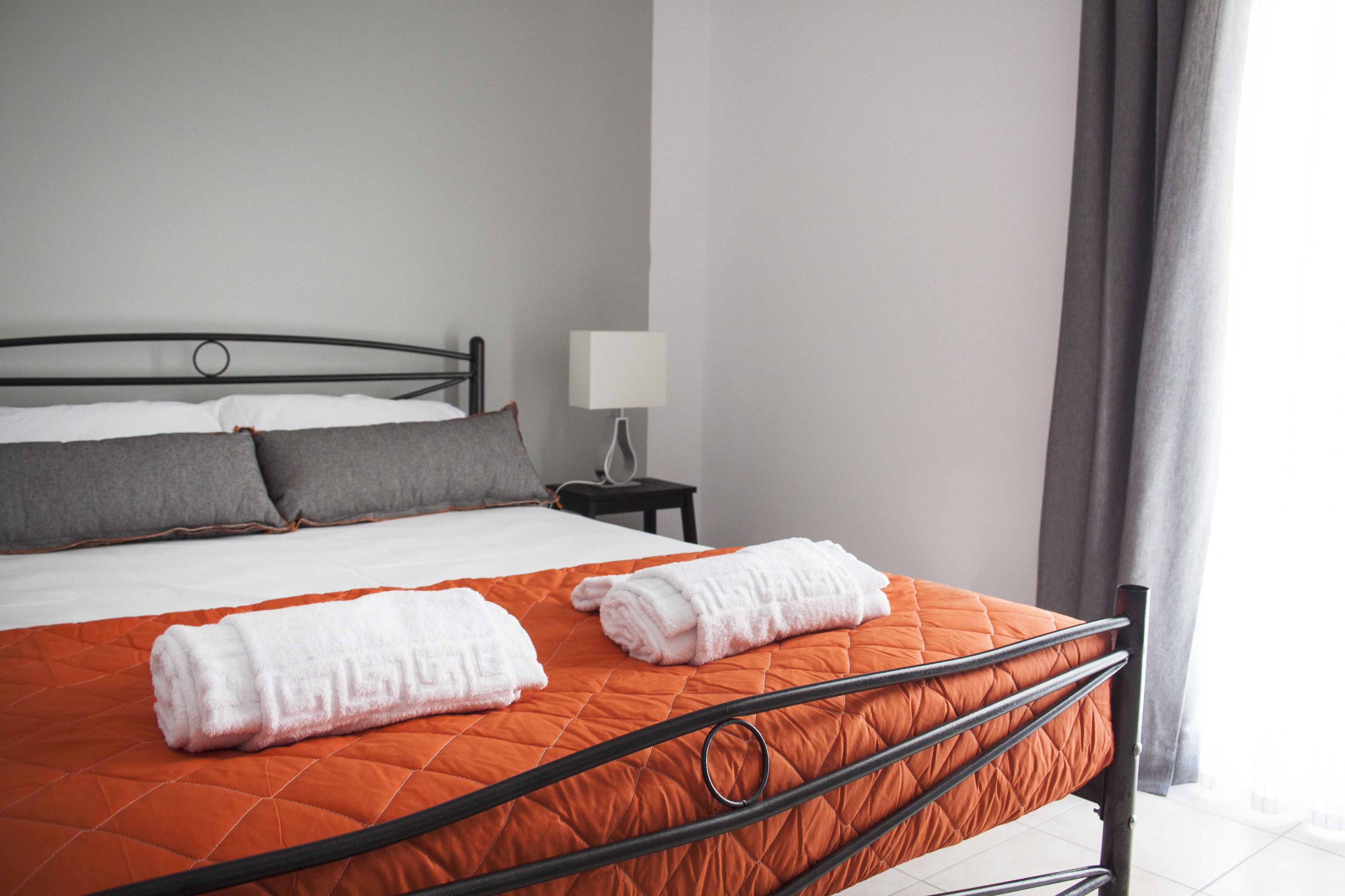 A neatly made bed with an orange quilt, white pillows, and two rolled white towels.