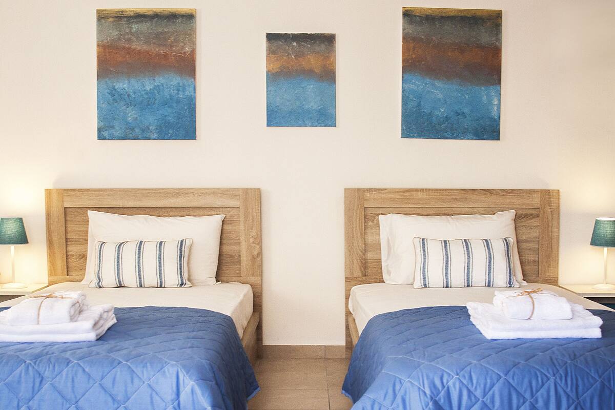 A hotel room with two beds, each with a blue quilted bedspread and wooden headboards. There are three paintings on the wall above the beds, and a pair of lamps on either side of the room.
