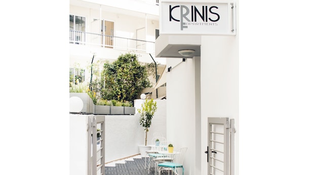 A narrow outdoor corridor with a white wall, a sign reading "KRINS APARTMENTS", and a small patio area with a table and chairs. - Krinis Apartments (Rhodes)