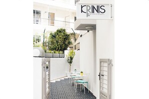 A narrow outdoor corridor with a white wall, a sign reading "KRINS APARTMENTS", and a small patio area with a table and chairs. - Krinis Apartments (Rhodes)