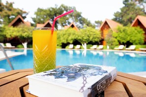 A glass of orange juice with a straw, a book titled 'CECTPA,' and a pool with lounge chairs and wooden cabins in the background. - Woodline Hotel (Kemer)