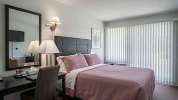 A hotel room with a bed, desk, chair, lamp, and a large window with blinds.