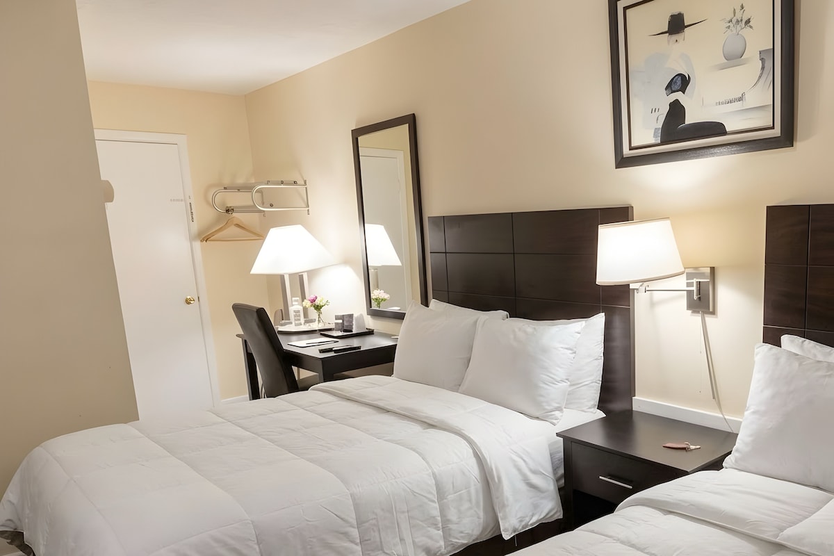 A hotel room with two beds, a desk, a chair, a lamp, and a framed picture on the wall.