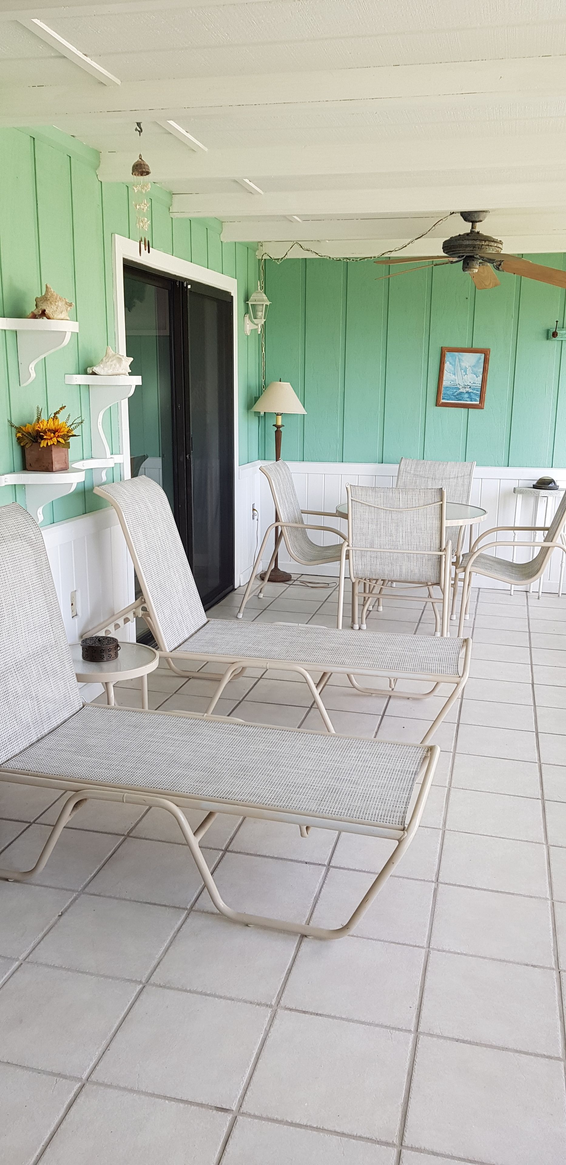 Een overdekt terras met rieten stoelen en tafels, een plafondventilator en een deur naar binnen.