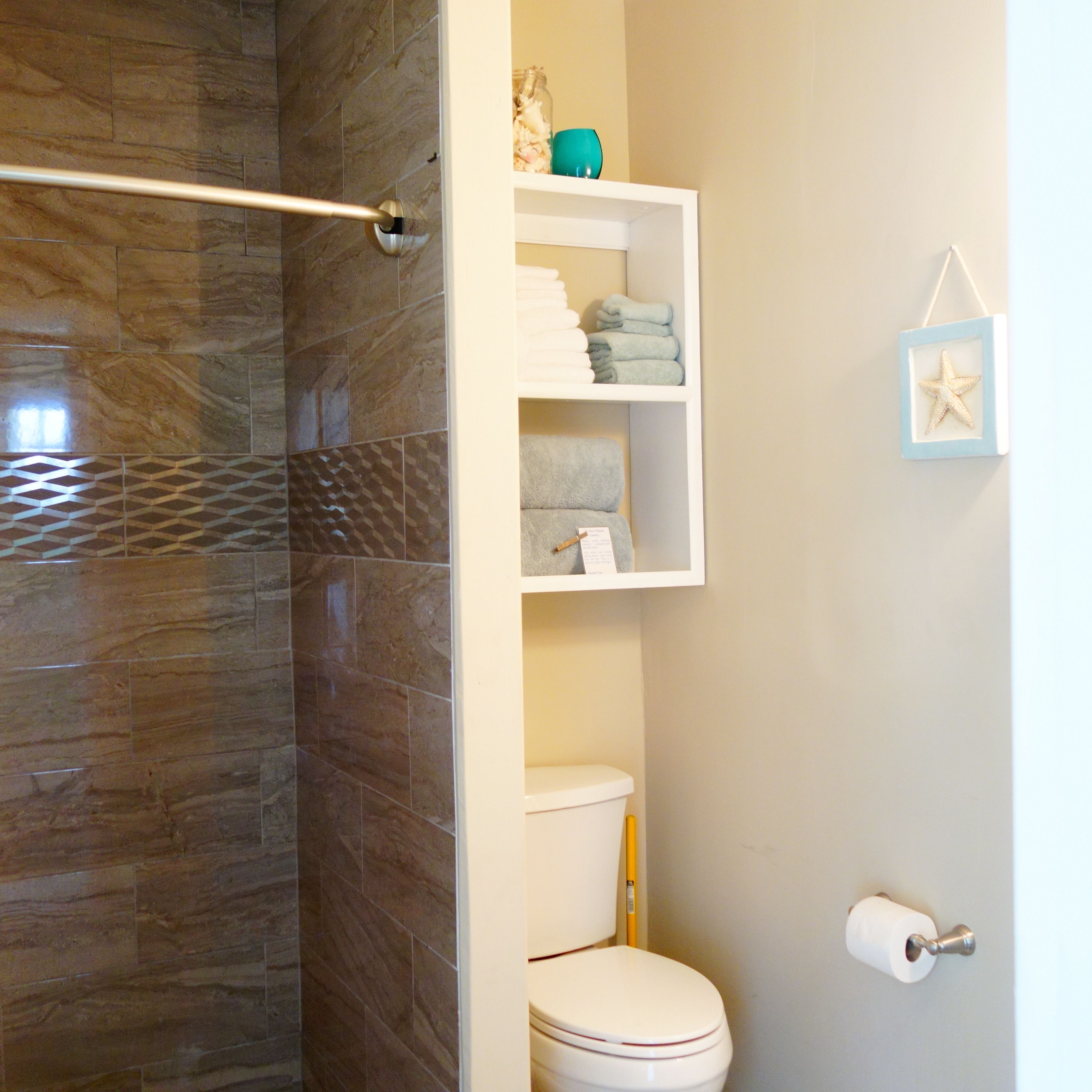 Un baño con pared de ducha de mármol, inodoro y repisa con toallas dobladas y artículos de tocador.