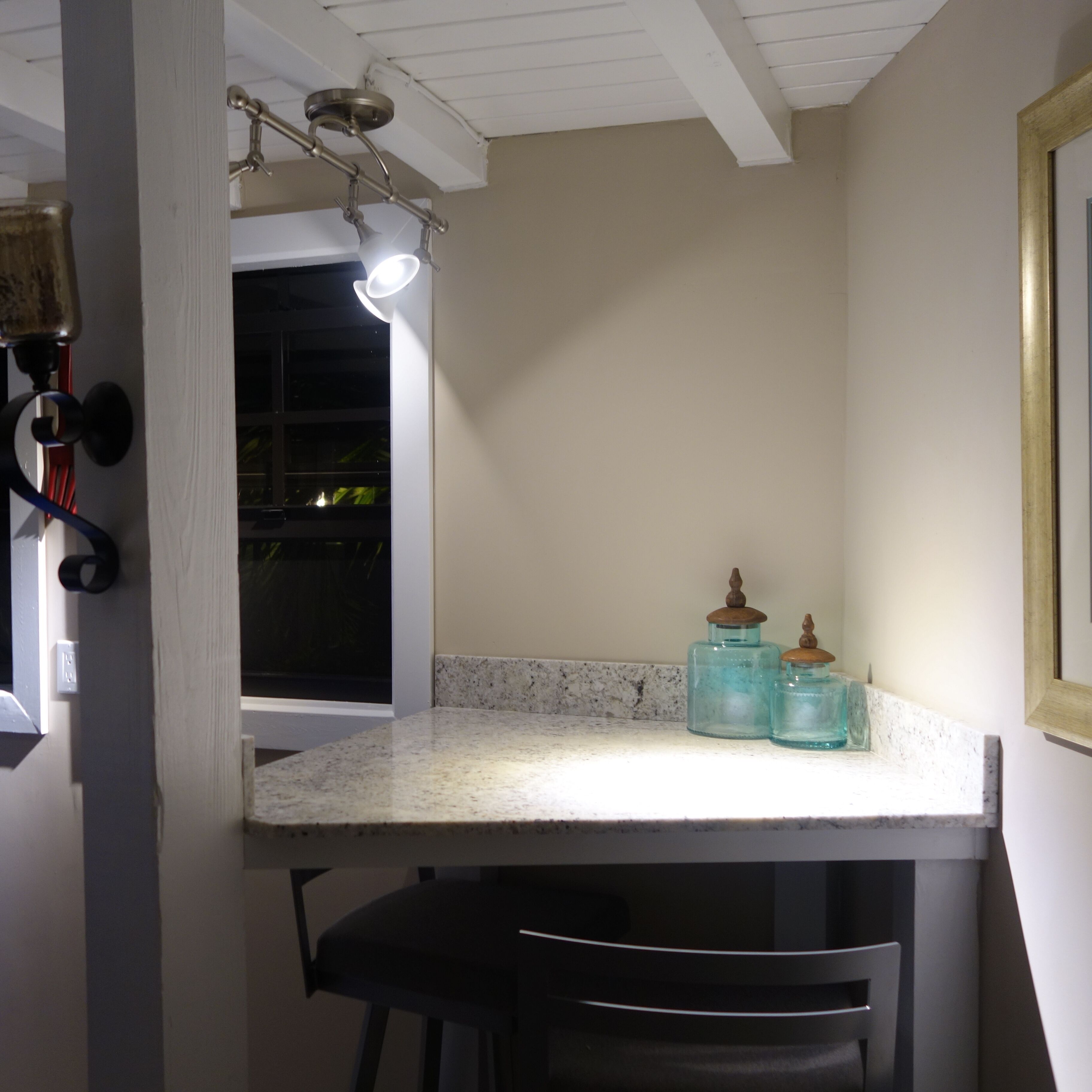 A kitchen with a white countertop, two glass jars with wooden lids, a black chair, and a window with a view of greenery.