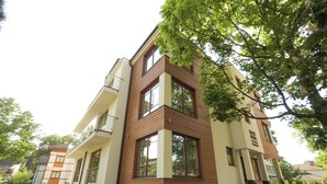 Un edificio moderno a più piani con pannelli esterni misti in legno e beige, ampie finestre e un cortile pavimentato.