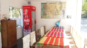 A room with a dining table covered with a colorful tablecloth, wooden chairs, a globe on the table, a Coca-Cola refrigerator, and a map on the wall. - Hostel Roma Inn 2 (Kalaw)
