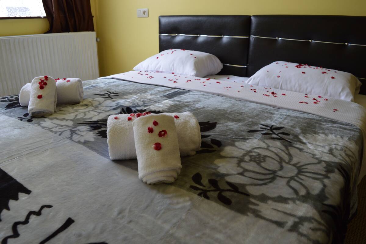 A neatly made bed with floral bedding and a headboard, towels with red flowers, and a window with curtains.