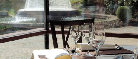A table set for dining with a book, glasses, and cutlery, overlooking a fountain and greenery.
