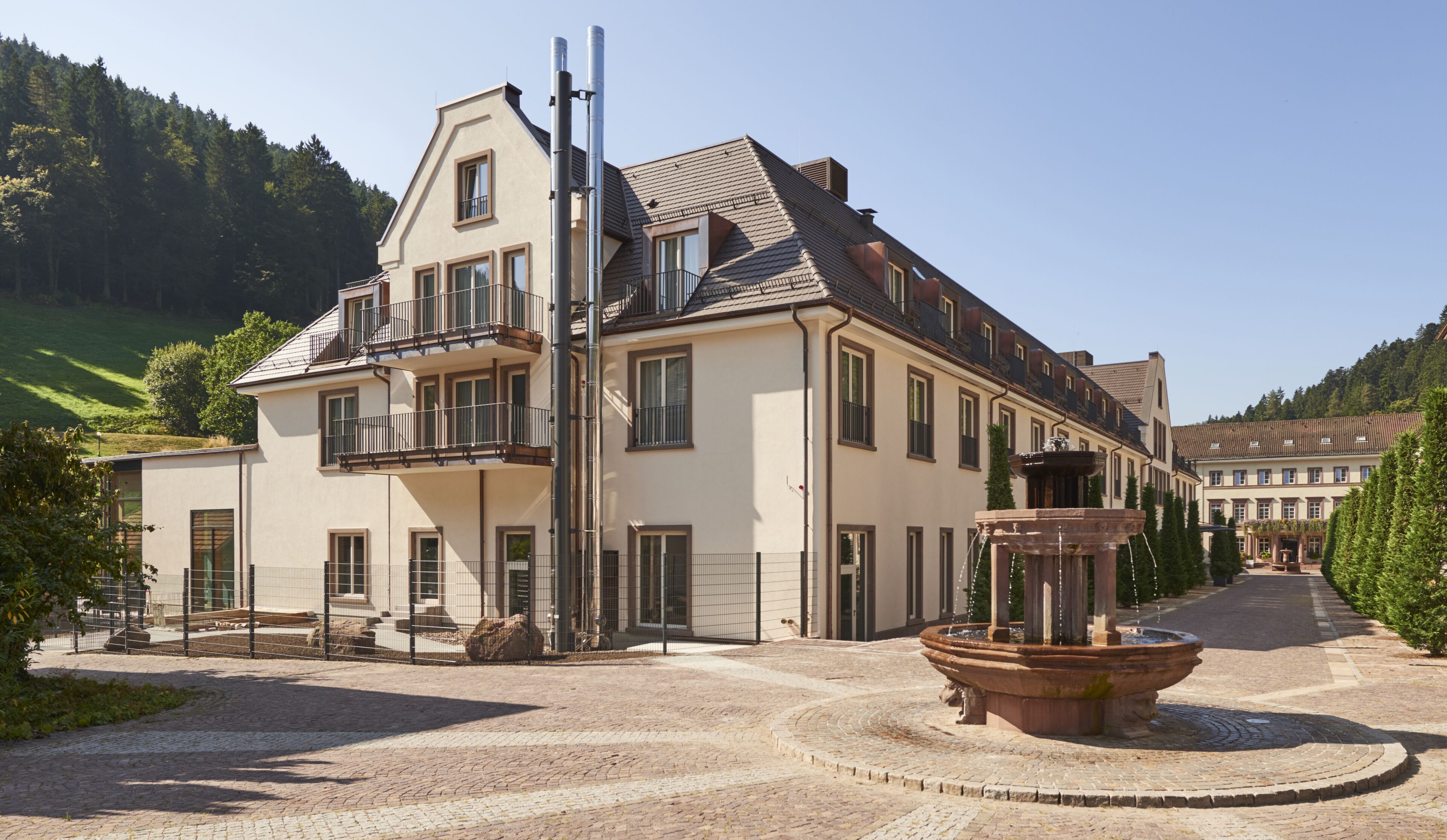 A large, multi-story building with a prominent balcony, a fountain in the courtyard, and a hilly landscape in the background.
