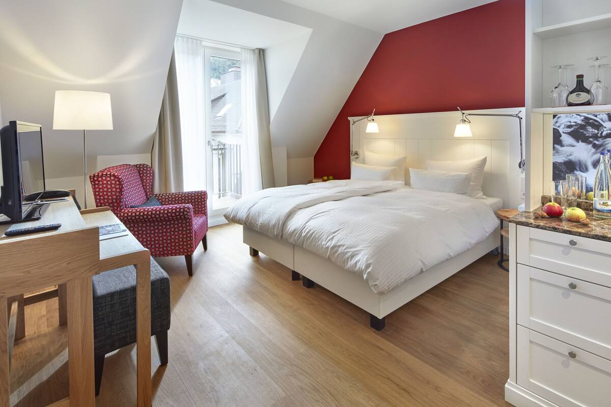 A hotel room with a bed, desk, chair, and a red accent wall.