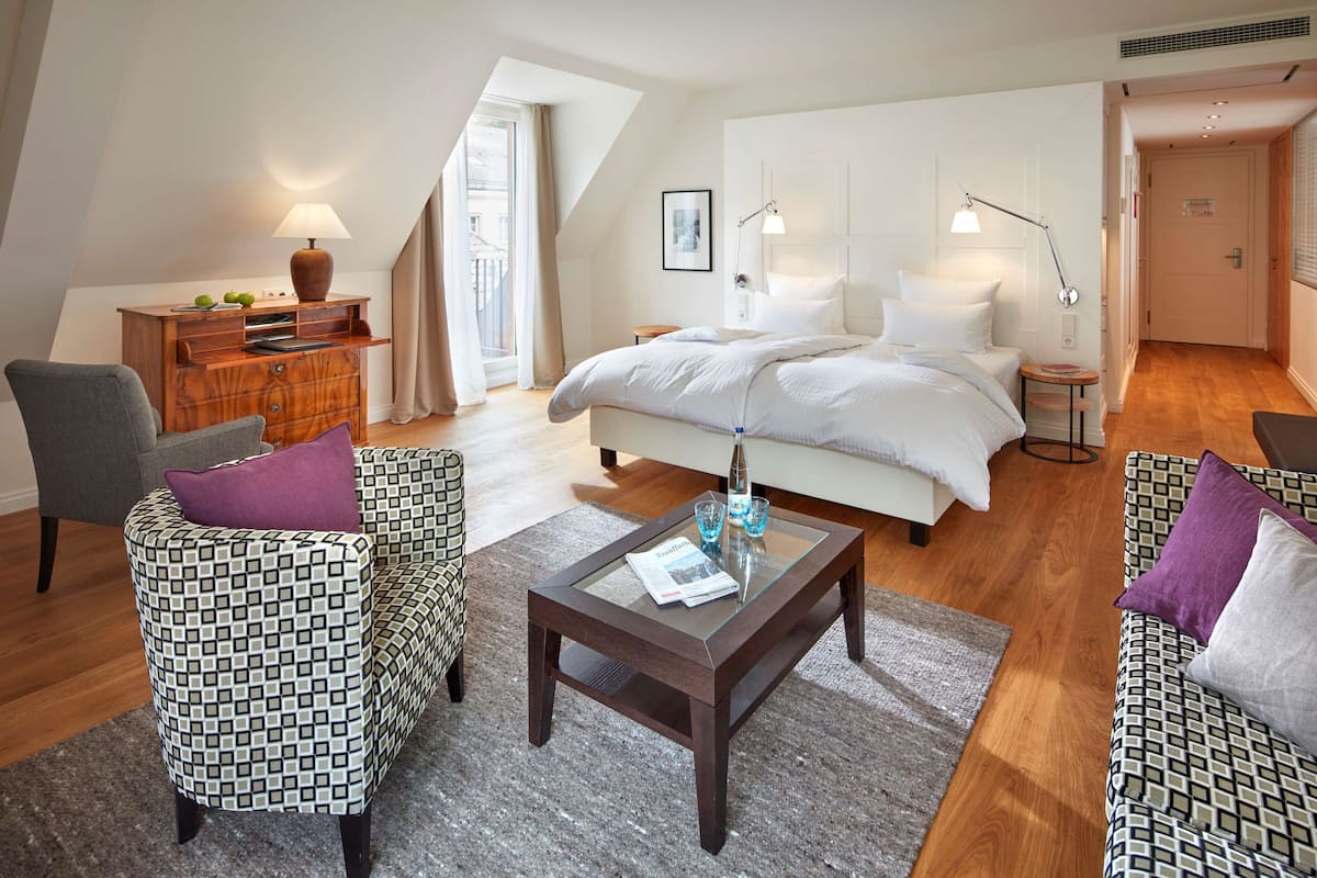 A modern hotel room with a large bed, two armchairs, a wooden side table, a glass coffee table, and a small round table with a lamp.