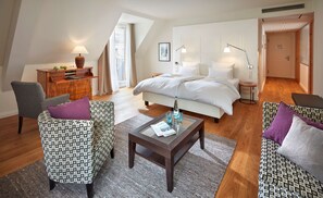 Una habitación de hotel moderna con una cama grande, dos sillones, una mesita de madera, una mesa de centro de cristal y una pequeña mesa redonda con una lámpara.