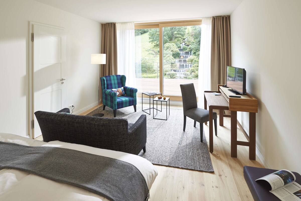 A hotel room with a bed, a desk with a computer, a chair, a sofa, and a view of a waterfall.
