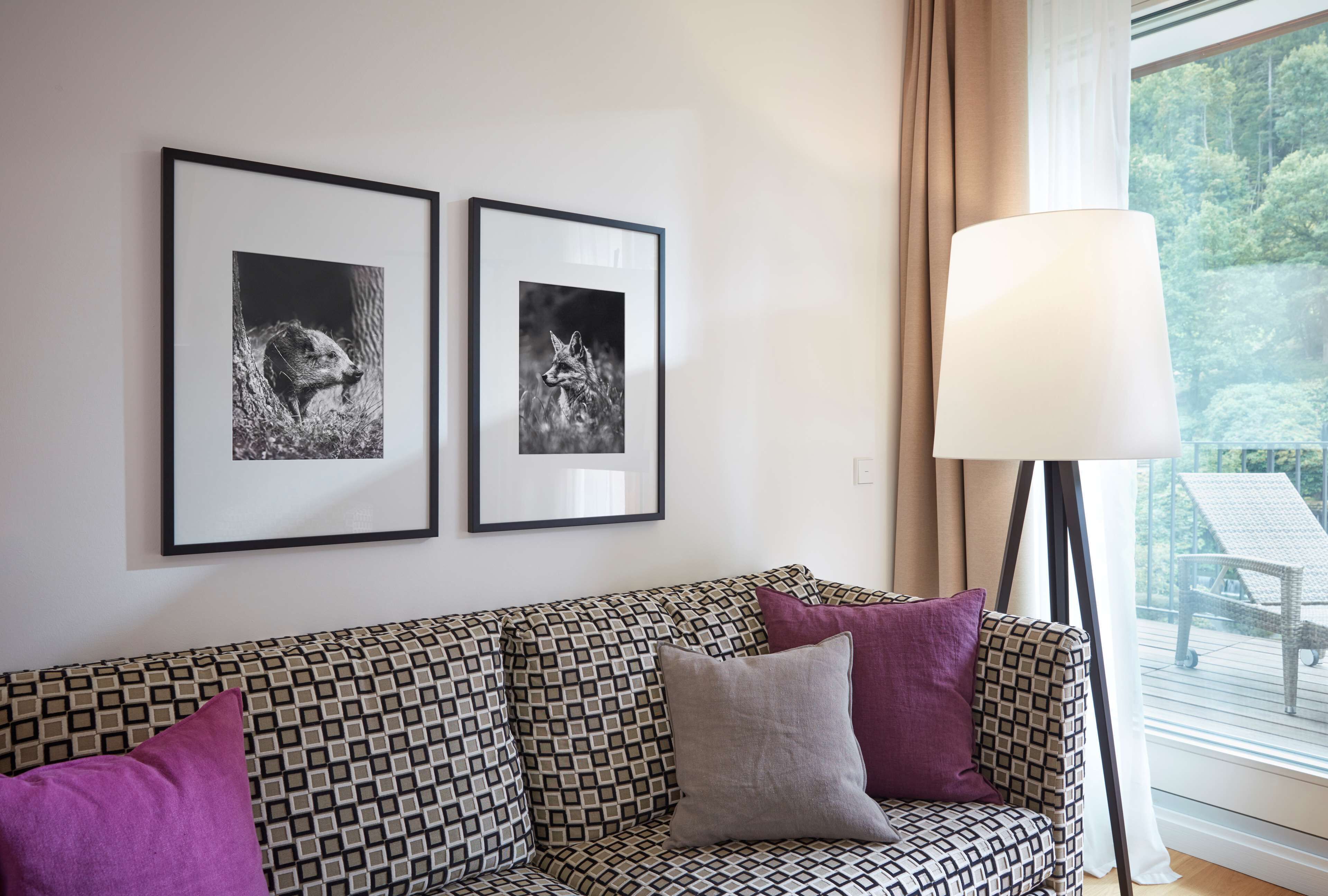 Een gestreept bankstel met twee ingelijste foto's van dieren aan de muur, een vloerlamp en uitzicht op een terras door een raam.