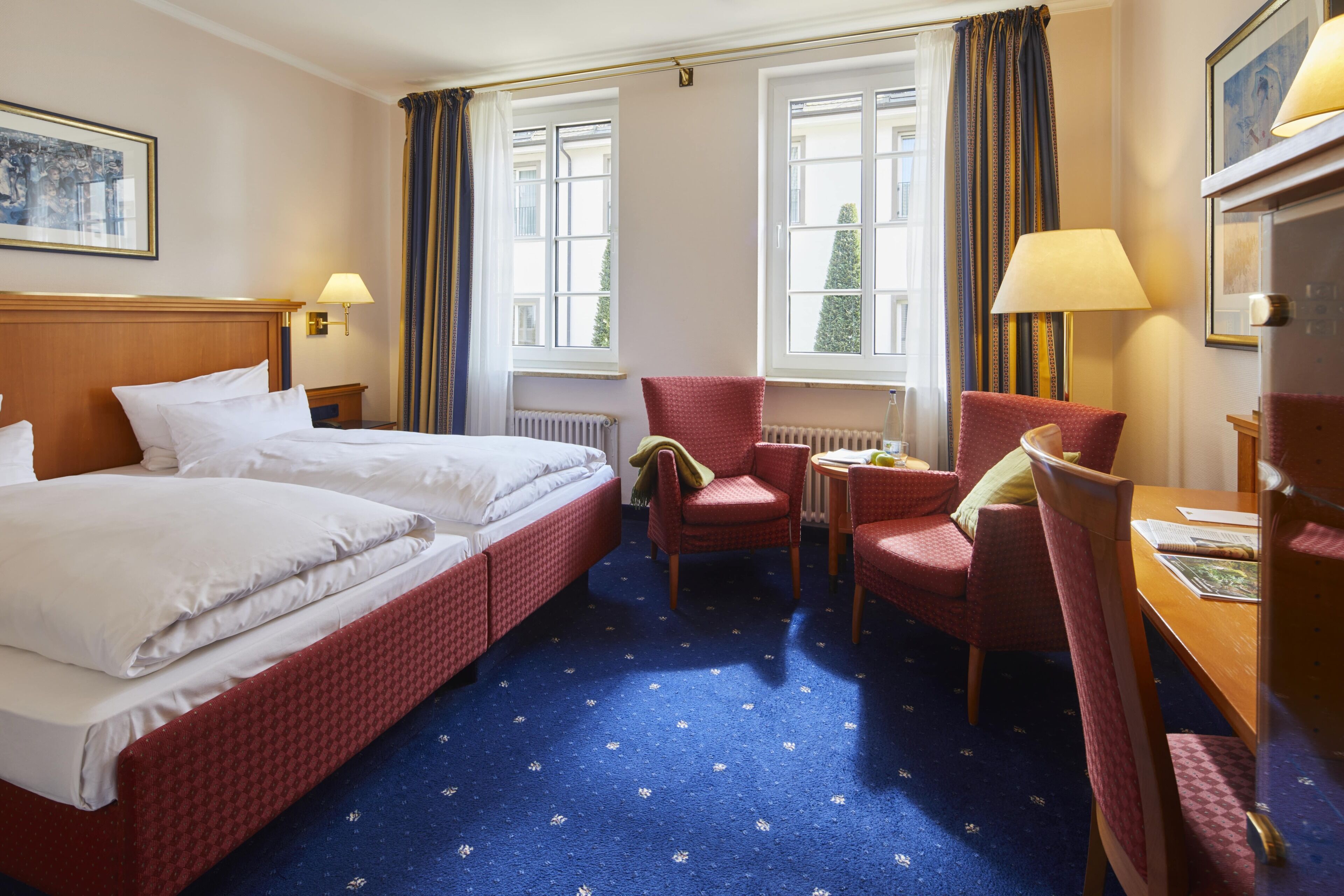 Habitación de hotel con cama doble, dos sillas, una mesita y dos ventanas con cortinas.