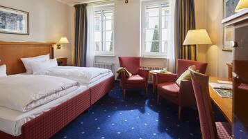 Habitación de hotel con cama doble, dos sillas, una mesita y dos ventanas con cortinas.