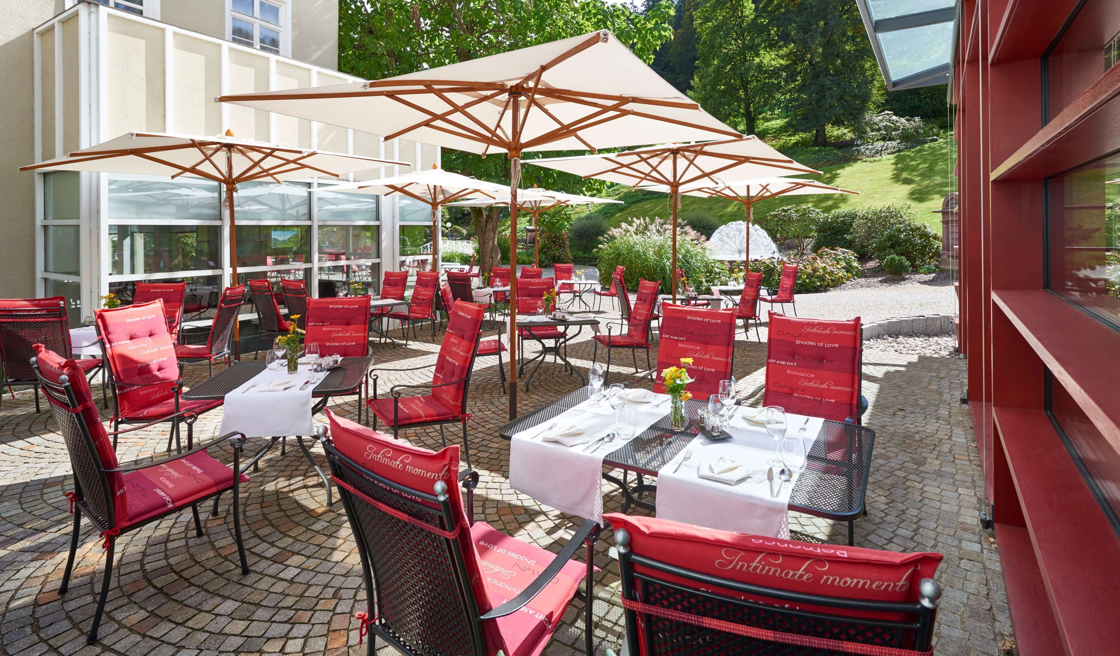 Un área de comedor al aire libre con sillas rojas y mesas blancas, cada una con mantel blanco y un atril para menús.