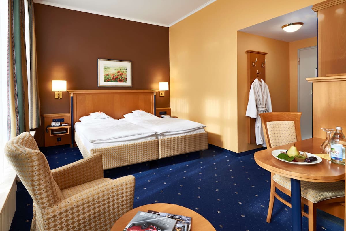 A hotel room with a bed, two bedside tables, a chair, a table with a plate of fruit and a bottle, a robe hanging on a hook, and a painting on the wall.