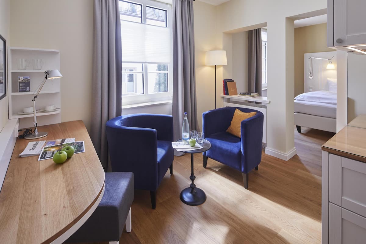 A hotel room with a wooden desk, two blue armchairs, a small round table with a lamp, a bed, and a window with curtains.