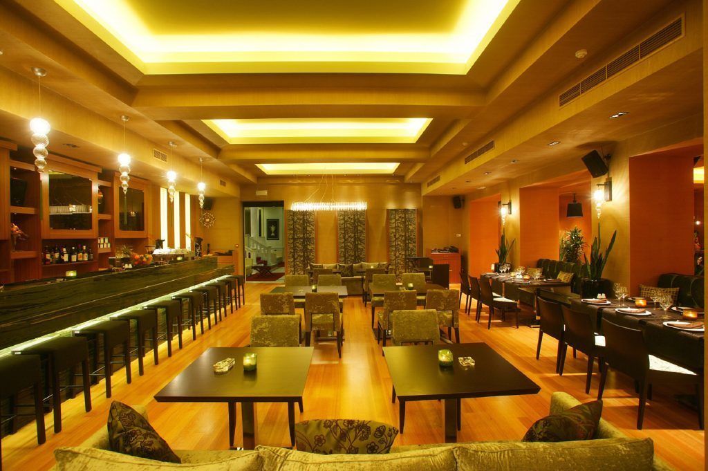 Un hall d’hôtel spacieux comprenant un bar, plusieurs espaces aménagés pour s’asseoir et un grand lustre central.
