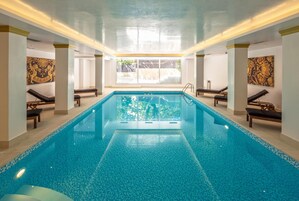 Una amplia piscina interior con agua cristalina y azul, rodeada de sillas de descanso y bancos, con grandes ventanales que dejan entrar luz natural.