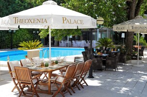 Un área de comedor al aire libre con mesas y sillas, una piscina y sombrillas con la marca "Theoxenia Palace".