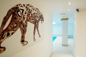 Un interior moderno con una escultura metálica geométrica en la pared, un suelo blanco impecable y una vista parcial de un área de piscina al fondo.
