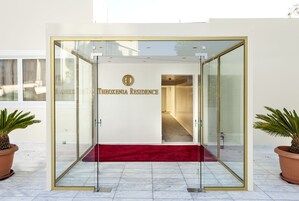 Entrada de un hotel con puerta de vidrio, alfombra roja y un letrero que dice 'Theoxenia Residence'.