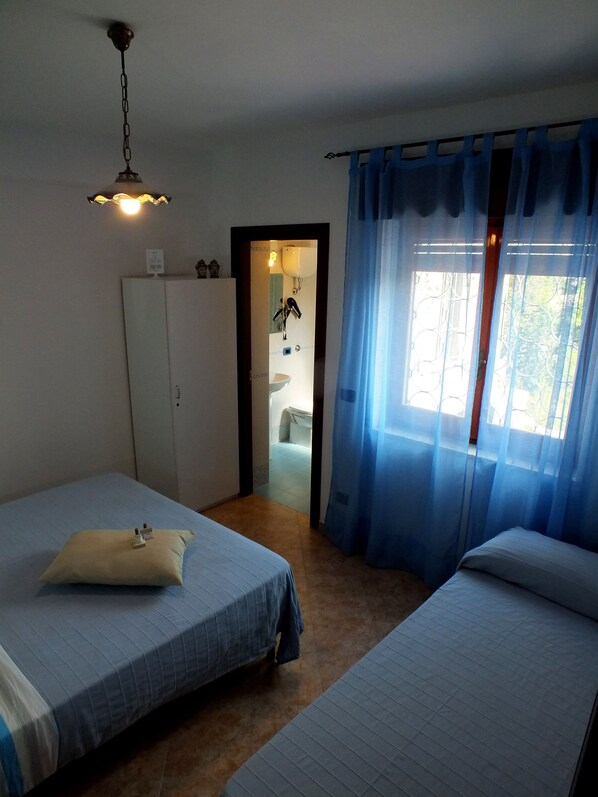 Una habitación con dos camas, un armario, un baño visible a través de una puerta y una ventana con cortinas azules.