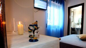 Une chambre d’hôtel avec un lit, une table de chevet ornée d’une bougie et d’une statuette, un téléviseur fixé au mur et une fenêtre avec des rideaux.