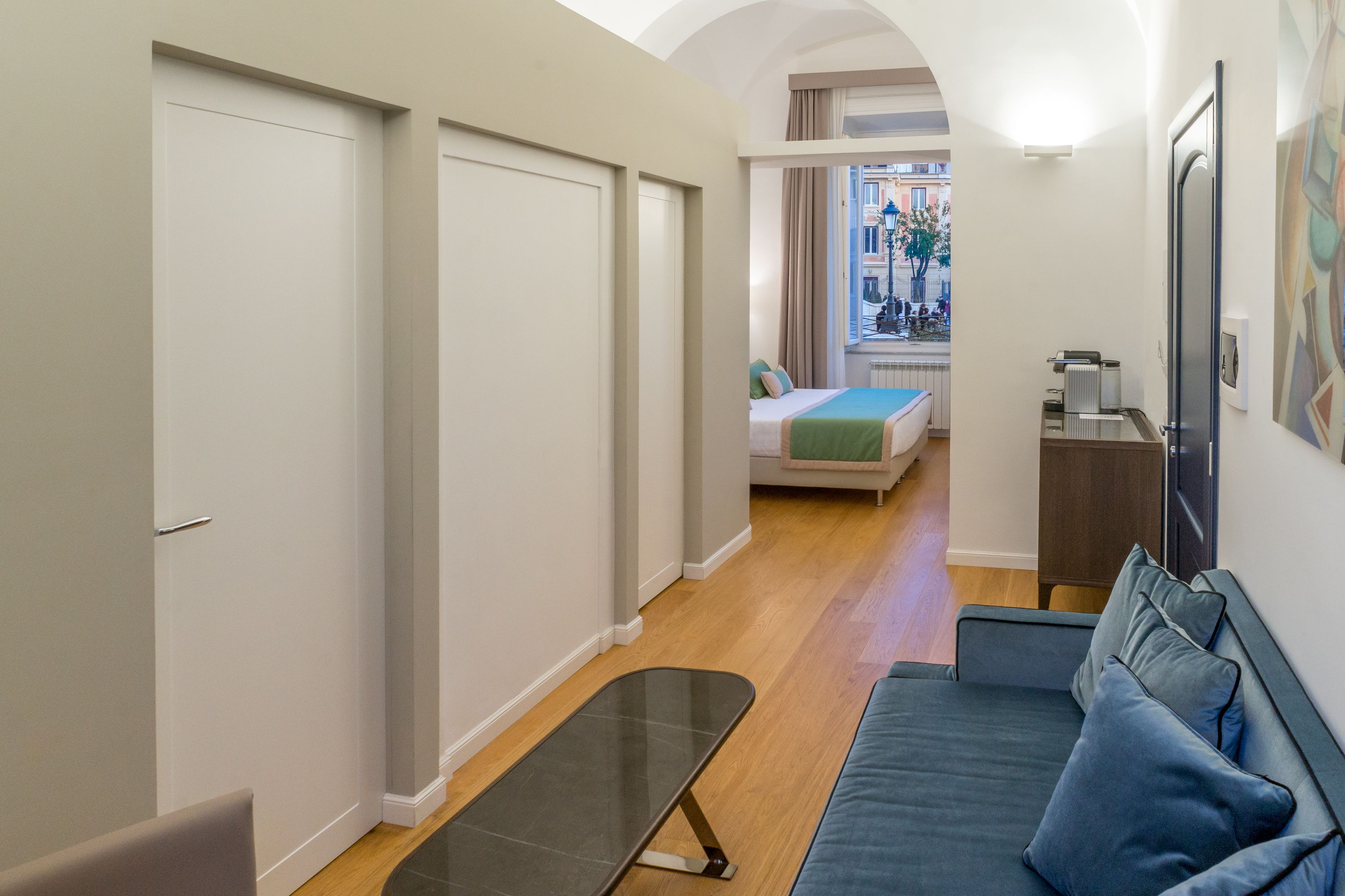 A modern hotel room with a sofa, a small table, and a view of the street through an open door.