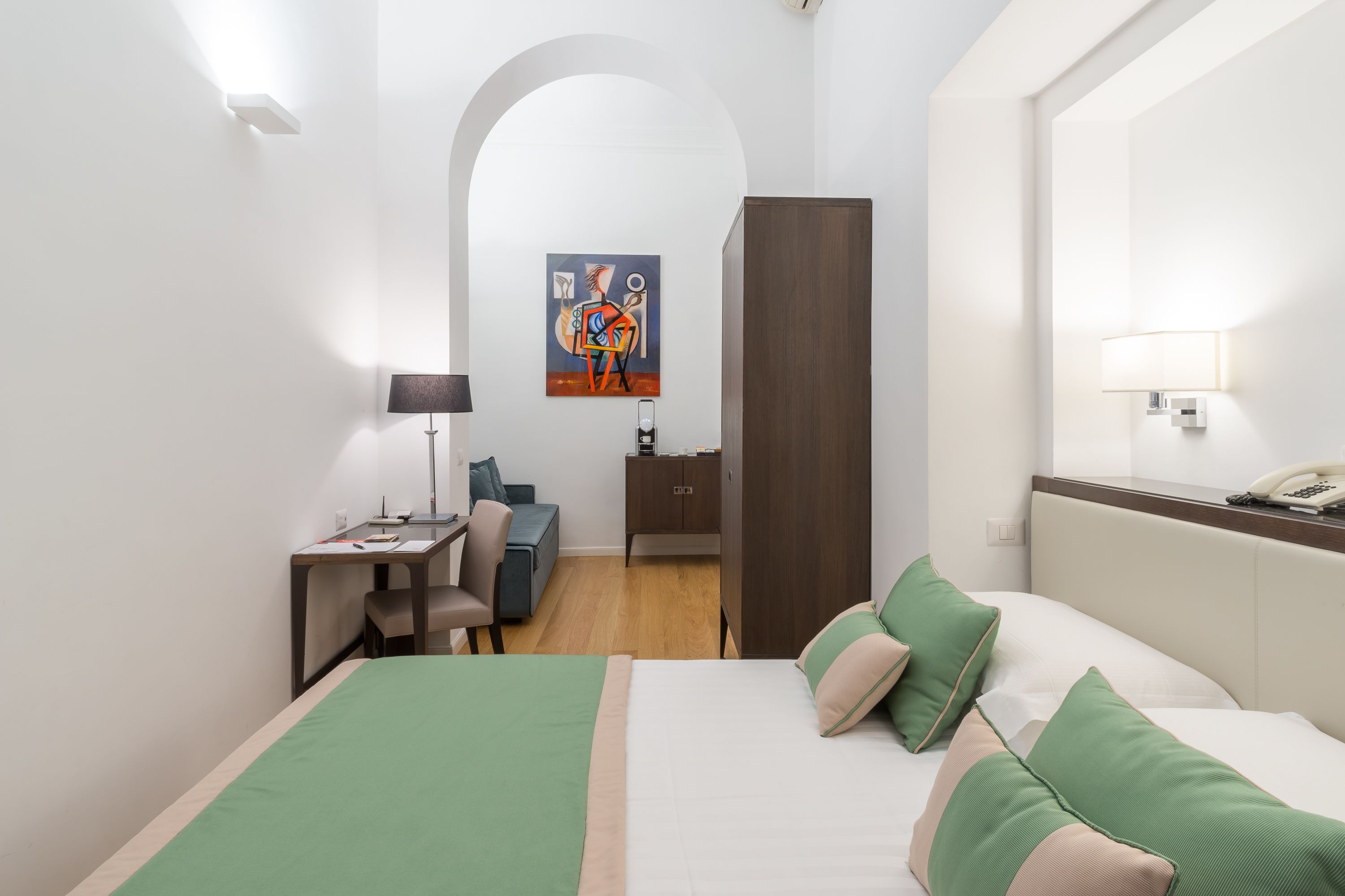 A hotel room with a bed, desk, chair, and a painting on the wall.