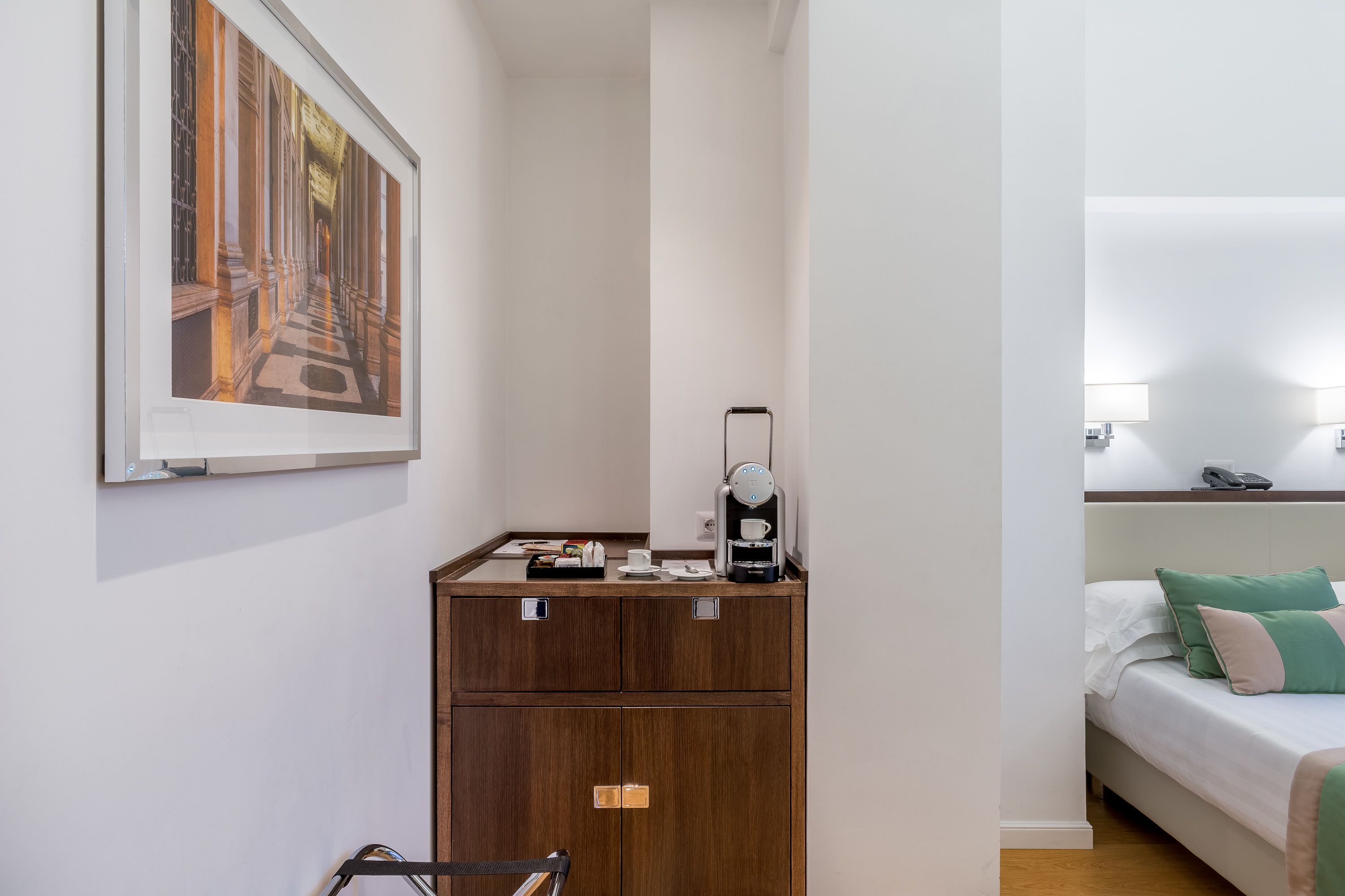 A hotel room with a bed, a nightstand, a coffee machine, a framed picture on the wall, and a wooden cabinet.