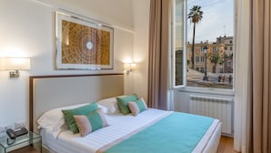Habitación de hotel con una cama grande, una mesita de noche de cristal, un teléfono, una obra de arte enmarcada sobre la cama y una ventana con vistas a edificios y una palmera.