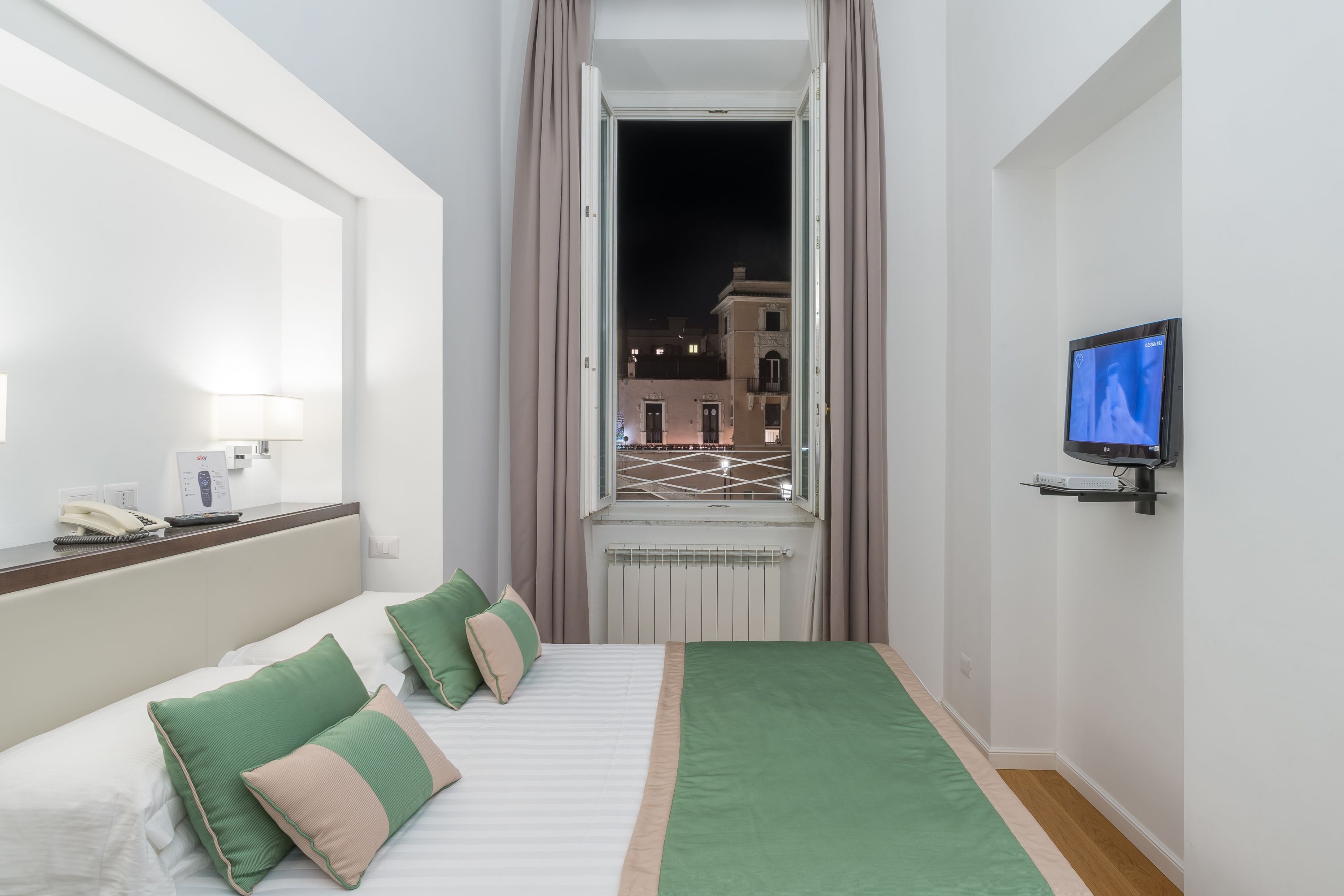 Habitació d'hotel amb un llit, un televisor fixat a la paret, una finestra amb cortines i vista a edificis.