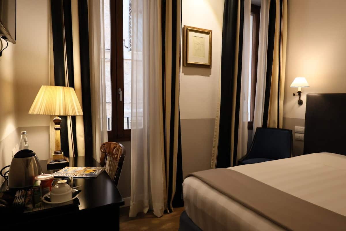 A hotel room with a bed, a desk with a lamp, a chair, and a window with curtains.