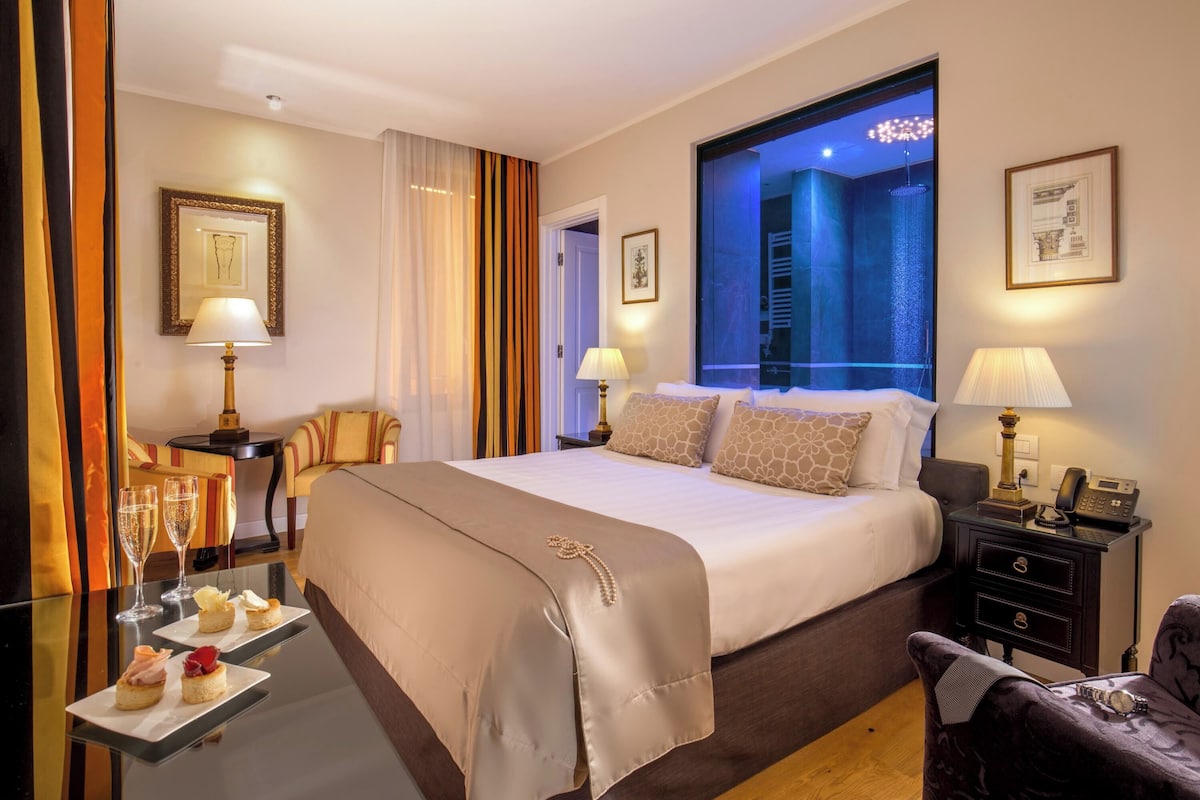 A hotel room with a large bed, bedside tables, a chair, and a view of a pool area.