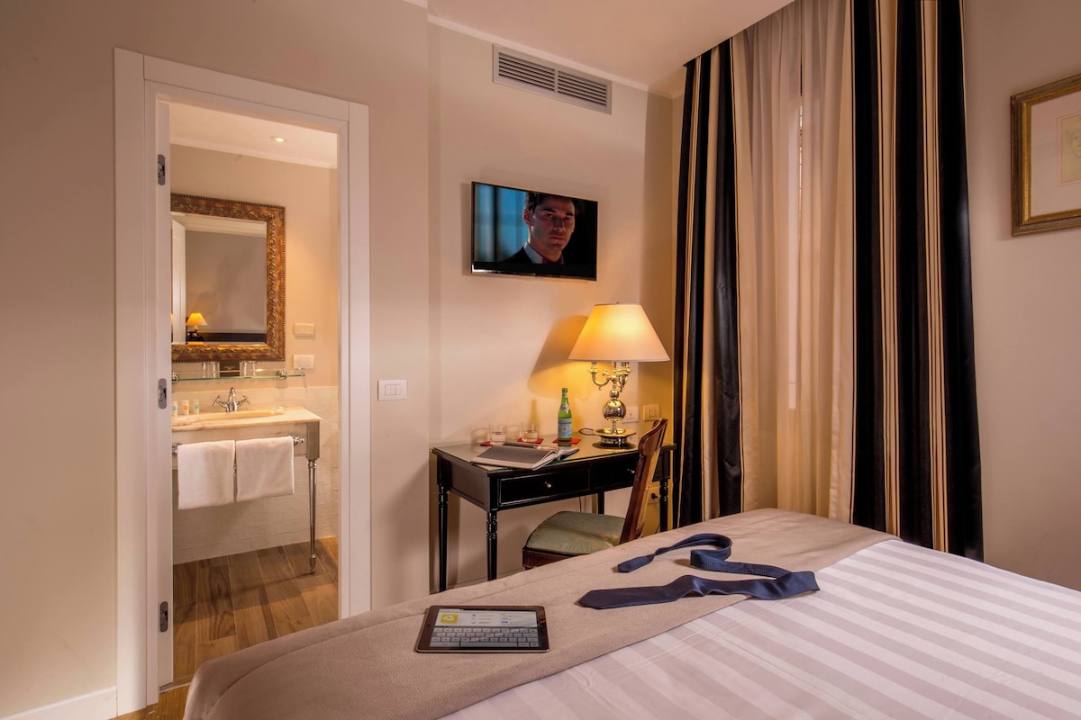 A hotel room with a bed, a desk with a lamp, a television, and a bathroom visible through an open door.