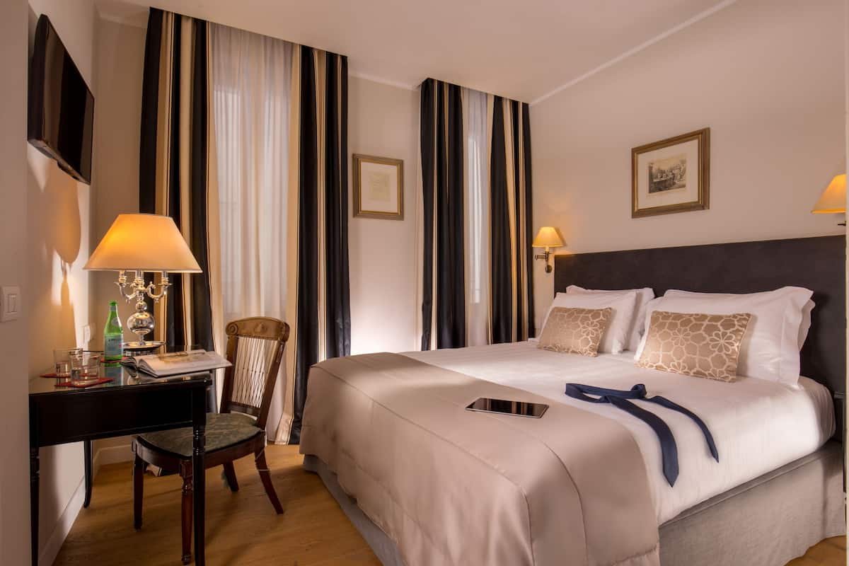 A hotel room with a bed, bedside lamps, a desk with a chair, a television, and a framed picture on the wall.