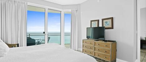 A hotel room with a large bed, a television on a wooden dresser, and a balcony with a view of the ocean.