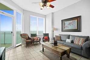 A living room with a sofa, two armchairs, a coffee table, and a ceiling fan. There is a large window with blinds, a door leading to a balcony, and a framed picture on the wall. - Aqua Beach Resort by Panhandle Getaways (Panama City Beach)